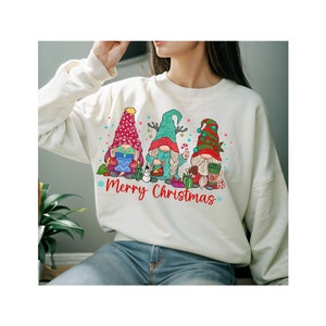 May include: Cream-colored sweatshirt featuring a festive design with three Christmas gnomes. The gnomes wear colorful hats and hold gifts and holiday treats. The words "Merry Christmas" are written in red script below the gnomes.