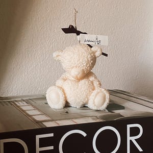 Può includere: Una candela a forma di orsetto color crema con superficie strutturata, appoggiata su un libro con la scritta "DECOR" in bianco. La candela ha un'etichetta con scritto "Wakino's Arts".