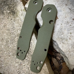 May include: Two green textured knife scales with rounded edges and multiple holes. The scales are made of a durable material and are designed to fit a knife handle.
