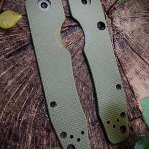 May include: Two green textured knife scales with rounded edges and multiple holes. The scales are made of a durable material and are designed to fit a knife handle.