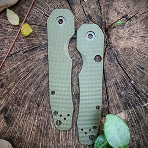 May include: Two green knife scales with a textured surface and multiple holes. The scales are on a wooden surface.