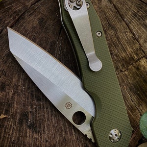May include: A green folding knife with a silver blade and a pocket clip. The knife has a textured green handle and a silver gear logo on the blade.