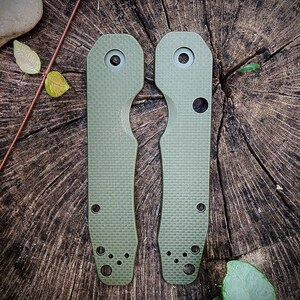 May include: Two green knife scales with a textured surface and multiple holes. The scales are on a wooden background.