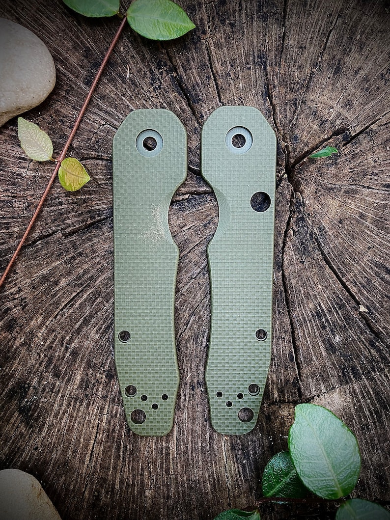 May include: Two green knife scales with a textured surface and multiple holes. The scales are on a wooden background.