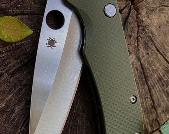 May include: A green folding knife with a partially open blade. The blade is made of stainless steel and has a spider logo on it. The handle is made of a textured green material and has a small silver button on it. The knife is sitting on a wooden surface.
