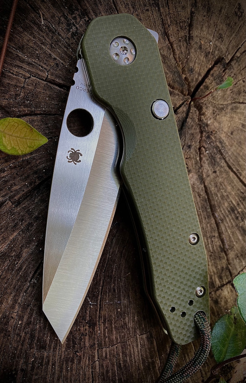 May include: A green folding knife with a partially open blade. The blade is made of stainless steel and has a spider logo on it. The handle is made of a textured green material and has a small silver button on it. The knife is sitting on a wooden surface.