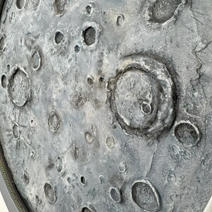 May include: A close-up of a gray and white textured surface with a circular shape. The surface has many craters and indentations, resembling the surface of the moon.