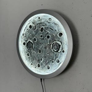 May include: A round, silver-colored wall lamp with a textured glass surface that resembles the moon. The lamp has a cord and plug.