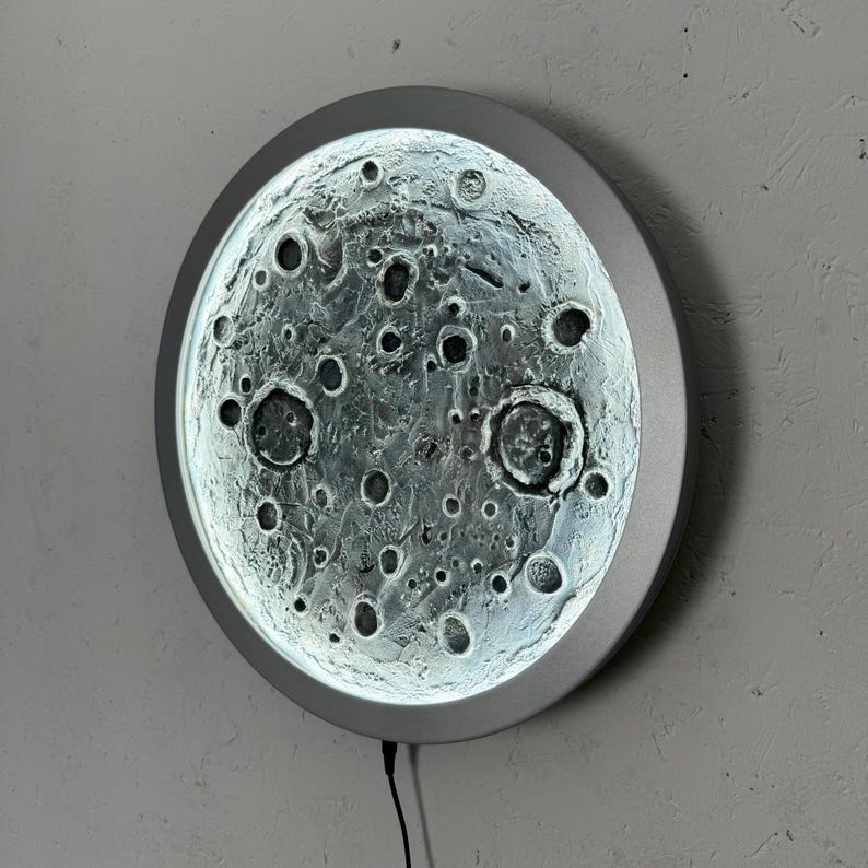 May include: A round, silver-colored wall lamp with a textured glass surface that resembles the moon. The lamp has a cord and plug.