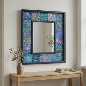 Op de afbeelding: Een vierkante wandspiegel met een donker frame en een rand bestaande uit individuele tegels in blauwe, paarse en gouden tinten. Elke tegel vertoont een uniek reliëfpatroon met bloemen of geometrische vormen, wat een textuurrijk mozaïekeffect creëert.