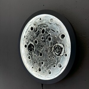May include: A white circular wall light with a 3D moon design. The moon has a textured surface with craters and shadows.