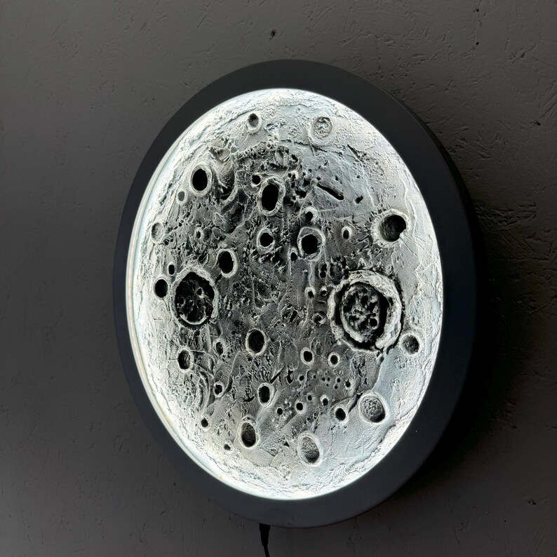 May include: A white circular wall light with a 3D moon design. The moon has a textured surface with craters and shadows.