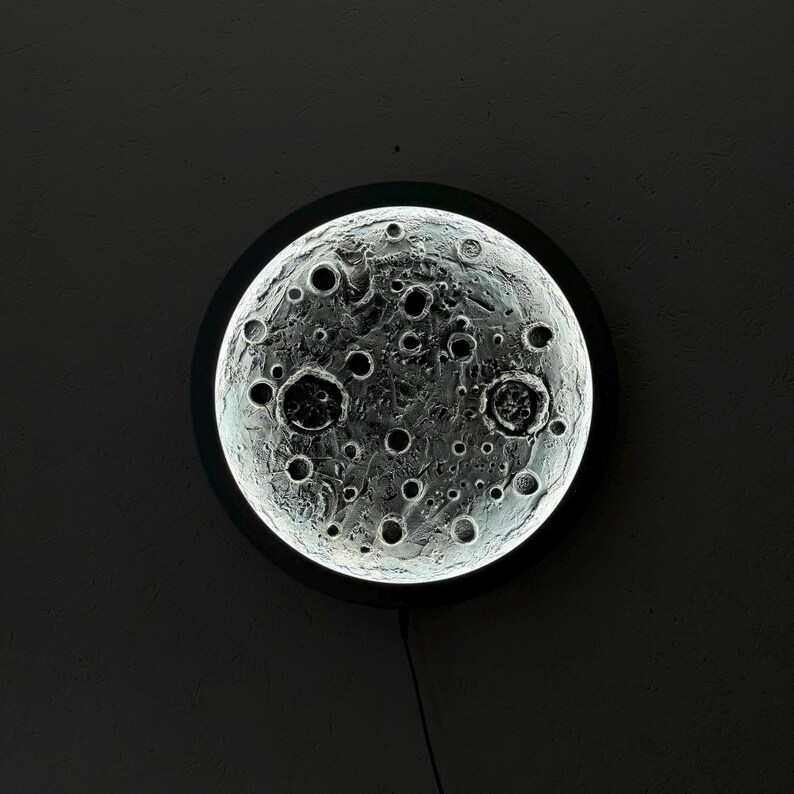 May include: A round, white moon-shaped light fixture with a black frame and a detailed, textured surface that resembles the moon's surface.