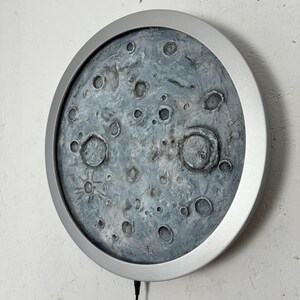 May include: A round, silver-framed wall decor featuring a textured, gray moon with craters. The moon is the focal point of the image, and the silver frame is a subtle accent.