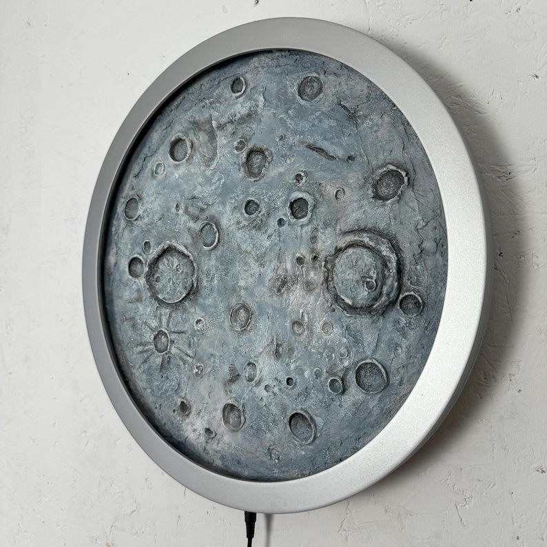 May include: A round, silver-framed wall decor featuring a textured, gray moon with craters. The moon is the focal point of the image, and the silver frame is a subtle accent.