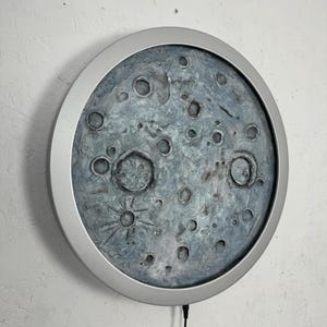May include: A round, silver-framed wall decor featuring a textured, gray and blue moon with craters. The moon is illuminated from behind, creating a soft glow.