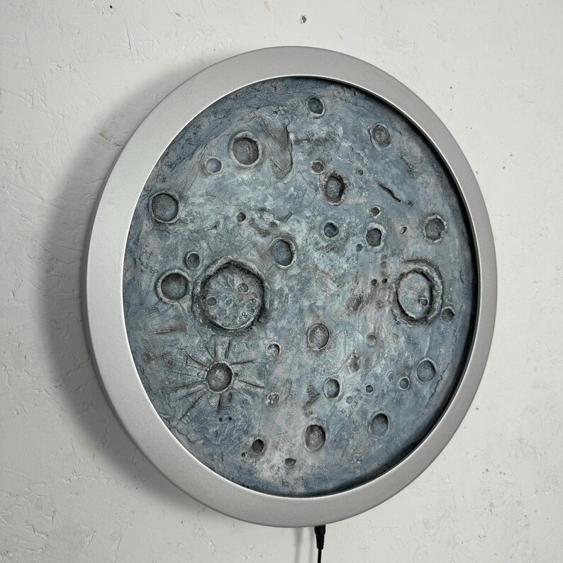 May include: A round, silver-framed wall decor featuring a textured, gray and blue moon with craters. The moon is illuminated from behind, creating a soft glow.