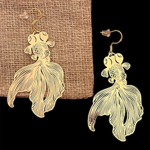 Puede incluir: Un par de pendientes de color dorado con forma de pez dorado. Los pendientes están detallados con intrincados recortes y tienen un diseño colgante.