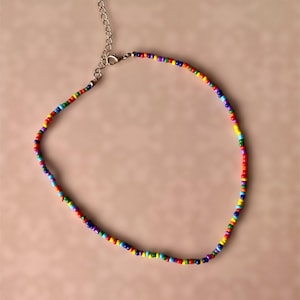 Peut inclure: Un collier de perles colorées avec une chaîne et un fermoir argentés. Le collier est composé de petites perles rondes aux couleurs de l'arc-en-ciel : rouge, orange, jaune, vert, bleu et violet. Le collier est présenté sur un fond rose clair.