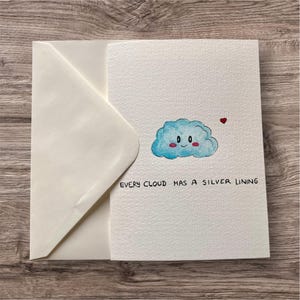 May include: A white greeting card with a blue cloud illustration and the text "EVERY CLOUD HAS A SILVER LINING." The card has a matching envelope. The cloud has a smiling face and a small red heart.