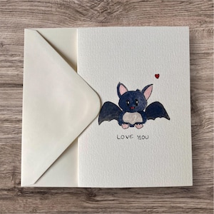 Peut inclure: Carte de vœux blanche avec une illustration à l'aquarelle d'une chauve-souris de dessin animé et un cœur rouge. Les mots "Love You" sont écrits sous la chauve-souris. Une enveloppe blanc cassé est à côté de la carte.