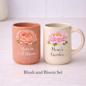 May include: A blush and bloom set of two ceramic mugs. One is peach-coloured with a peony and the text "Mom in Full Bloom." The other is cream-coloured with a pink peony and the text "Mom's Garden."