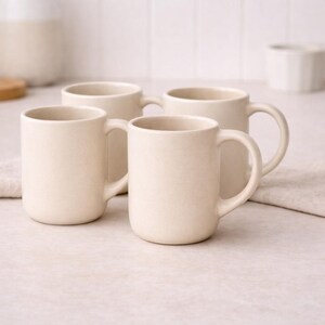 May include: Four cream-coloured ceramic mugs with rounded bodies and large handles are arranged on a light surface. The mugs are evenly spaced, suggesting a set. The background includes a white tiled wall and other neutral-toned kitchenware.