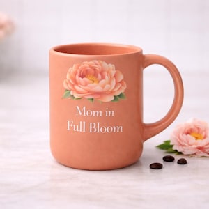 May include: A peach-coloured ceramic mug with a floral design and the text "Mom in Full Bloom." The mug has a curved handle and is accompanied by a flower and coffee beans. A vase of flowers is in the background.