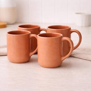 May include: Four terracotta-coloured ceramic mugs with rounded handles. The mugs are cylindrical and have a smooth, matte finish. They are arranged on a light-coloured surface, suggesting a kitchen or dining setting. The mugs are empty and ready for use.
