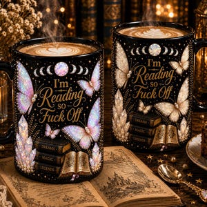May include: Two black ceramic mugs featuring a graphic of iridescent butterflies, crystals, and a stack of books labelled Dreams, Magic, and Legends. The text I'm Reading so F*** Off is printed in gold script. Each mug contains a hot beverage with latte art.