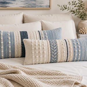 Puede incluir: Dos cojines lumbares rectangulares con un diseño texturizado de rayas verticales alternas en tonos azul, crema y blanco. La tela incorpora patrones tejidos, incluyendo filas de pompones, secciones trenzadas y bandas de color azul sólido.