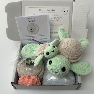 May include: Crochet kit for making two plush turtle toys. The kit includes yarn in green, beige, and pink, stuffing, safety eyes, a blunt needle, a stitch marker, and instructions. The kit is packaged in a white box with the words "Crochet Kit Turtles" on the front.