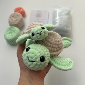 May include: A crocheted green turtle with a smaller crocheted beige turtle on its back. Both turtles have black eyes and a smiling mouth.