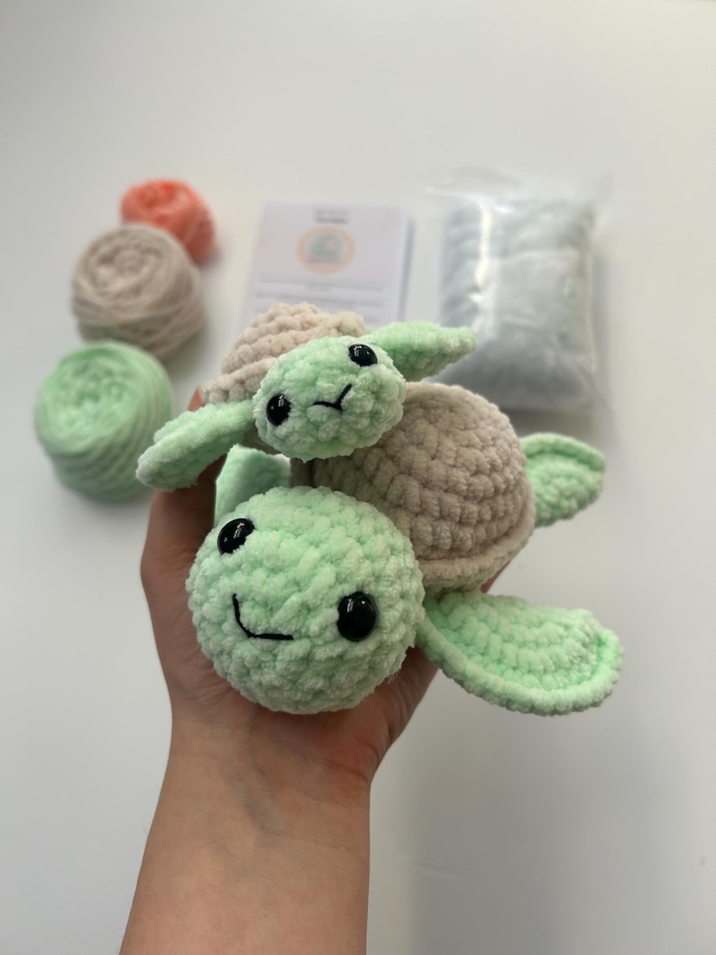 May include: A crocheted green turtle with a smaller crocheted beige turtle on its back. Both turtles have black eyes and a smiling mouth.