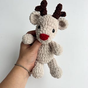 Op de afbeelding: Een handgemaakt, pluche rendier speelgoed in een lichtbeige kleur. Het heeft donkerbruine geweien, een rode neus en zwarte ogen. Het zachte, getextureerde materiaal geeft het een knuffelbaar uiterlijk, perfect voor kinderen of als vakantiedecoratie.