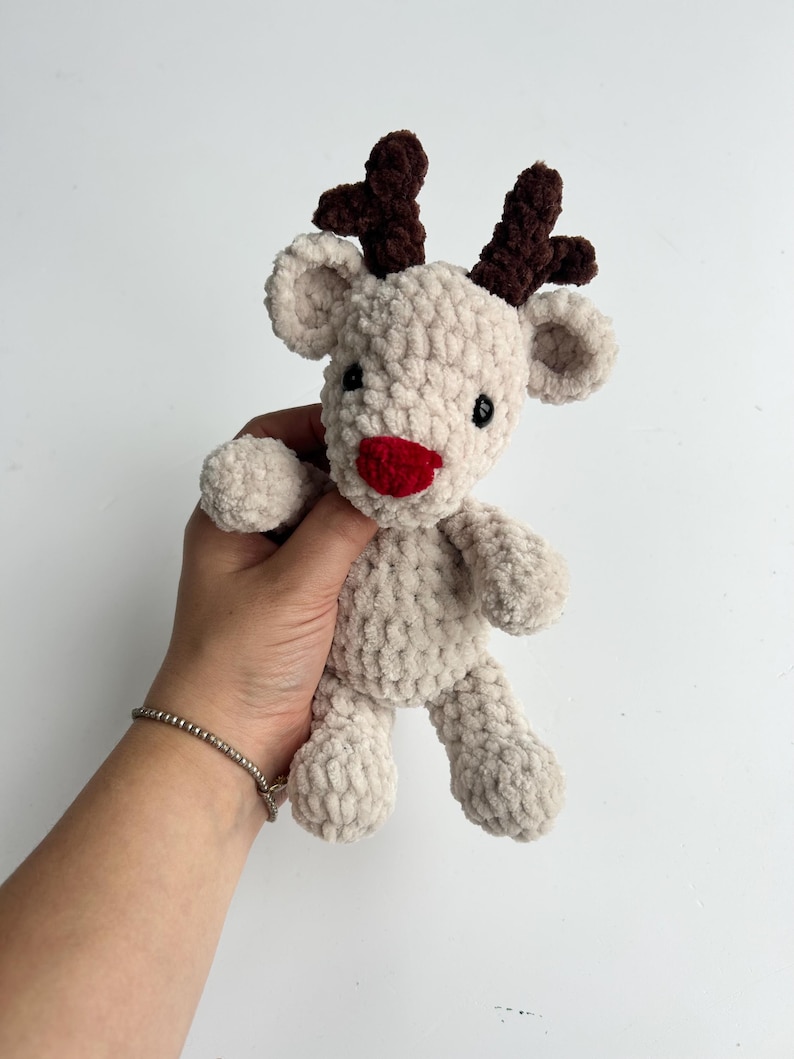 Op de afbeelding: Een handgemaakt, pluche rendier speelgoed in een lichtbeige kleur. Het heeft donkerbruine geweien, een rode neus en zwarte ogen. Het zachte, getextureerde materiaal geeft het een knuffelbaar uiterlijk, perfect voor kinderen of als vakantiedecoratie.