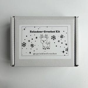 Op de afbeelding: Een witte rechthoekige doos met een label met de tekst "Reindeer Crochet Kit". Het label toont een cartoon rendier en sneeuwvlokken. De tekst "@sprinkleofcrochet" is ook zichtbaar.
