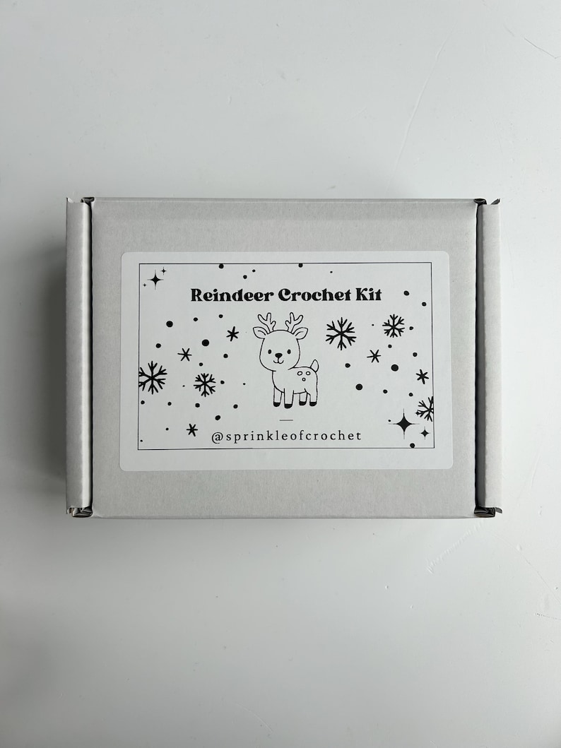 Op de afbeelding: Een witte rechthoekige doos met een label met de tekst "Reindeer Crochet Kit". Het label toont een cartoon rendier en sneeuwvlokken. De tekst "@sprinkleofcrochet" is ook zichtbaar.
