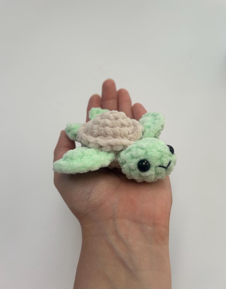 May include: A small crocheted sea turtle toy with a light green shell and a light pink underside. The turtle has black eyes and a small smile.