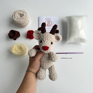 Op de afbeelding: Een beige gehaakt rendier met bruine geweien en een rode neus. De afbeelding toont ook beige en gele wol, een haaknaald, een naald en een zakje vulling. Een boekje met de titel "Reindeer" is ook zichtbaar.