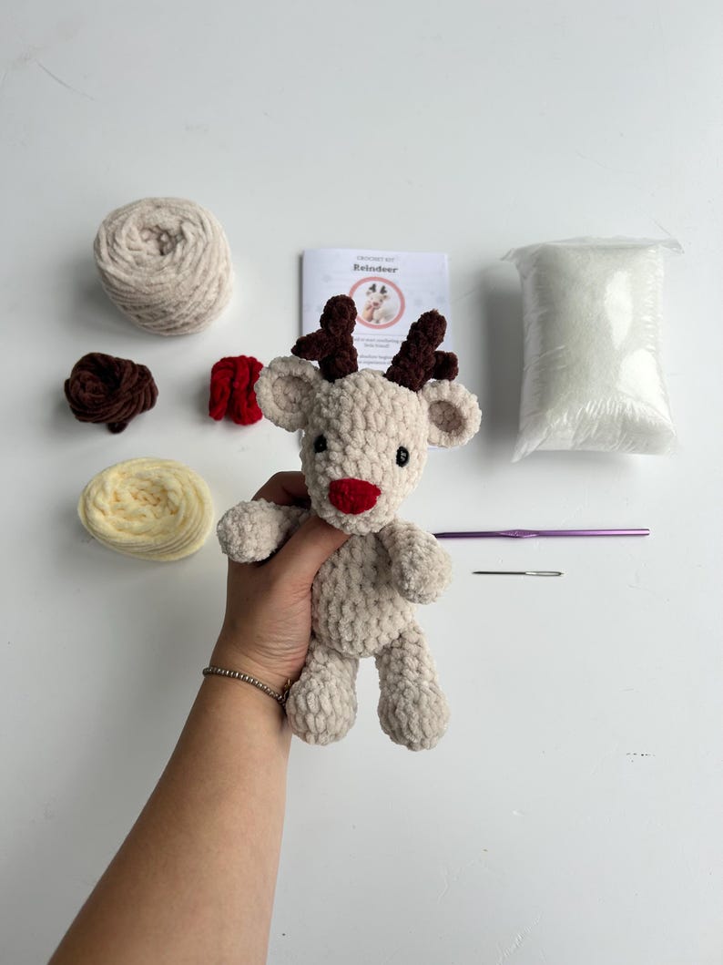 Op de afbeelding: Een beige gehaakt rendier met bruine geweien en een rode neus. De afbeelding toont ook beige en gele wol, een haaknaald, een naald en een zakje vulling. Een boekje met de titel "Reindeer" is ook zichtbaar.
