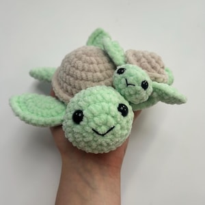May include: A crocheted green and pink turtle toy with a smaller green turtle on its back. The turtles have black eyes and a smiling mouth.