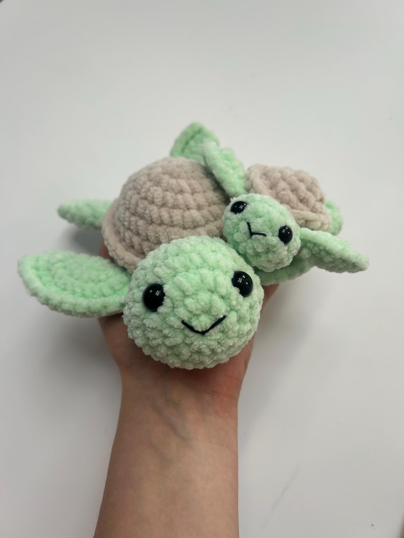 May include: A crocheted green and pink turtle toy with a smaller green turtle on its back. The turtles have black eyes and a smiling mouth.