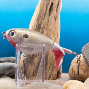 May include: A silver and red fishing lure with a fish-like design. The lure has a large eye, a black spot, and two treble hooks. The lure is displayed on a clear stand against a blue background.