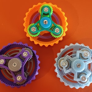 May include: Three colourful fidget spinners with gears. The spinners are purple, orange and light blue. Each spinner has a three-pronged centre with a small gear on each prong. The gears are connected to larger gears around the outside of the spinners.
