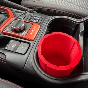 May include: A bright red cylindrical cup holder insert with vertical ridges along the interior walls, positioned inside the black plastic cup holder of a vehicle center console featuring a red trim panel labeled X-MODE and various control buttons.