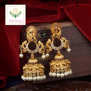May include: Gold earrings with a peacock design, white stones, and pearl accents. The earrings have a dangling bell-shaped charm.
