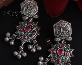 May include: A pair of silver earrings with intricate designs and red and green gemstones. The earrings have a hexagonal top with a floral pattern and dangle with multiple small bells.
