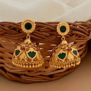 May include: A pair of gold-colored, bell-shaped earrings. Each earring features a green gemstone at the top and green and red accents on the bell. The earrings are intricately designed with floral and geometric patterns, and are displayed in a woven basket.
