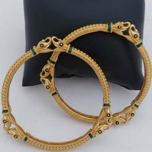 Könnte beinhalten: Zwei goldene Armreifen mit komplizierten Designs und grünen Emaille-Akzenten. Die Armreifen sind übereinander gestapelt und haben an den Enden ein vogelartiges Design.
